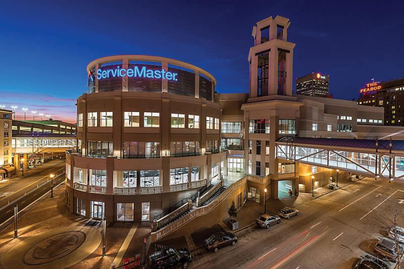 A modern, multi-story ServiceMaster building is lit up at dusk, with large windows and a skywalk connecting to another structure. City streets and distant illuminated signs are visible under a deep blue sky.