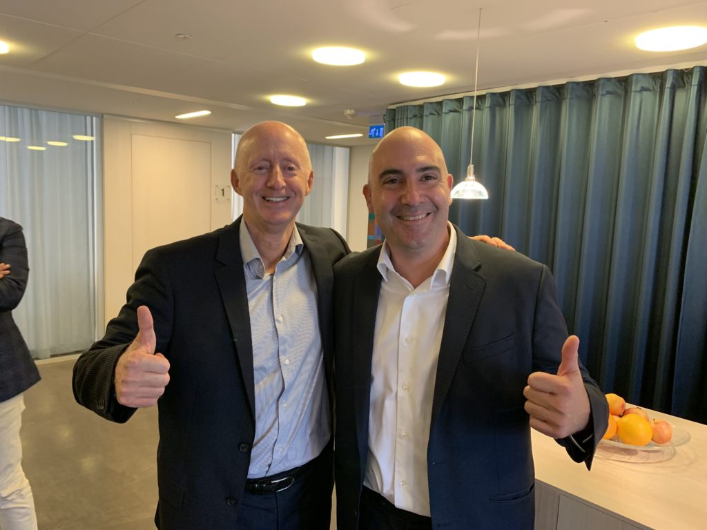 Two men in suits smile and give thumbs up while standing in a modern office space with blue curtains, pendant lights, and a table with fruit in the background.