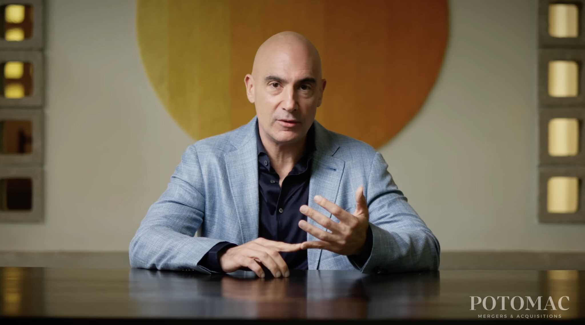 A man in a light blue blazer gestures with his hands while speaking at a table. The background features a large, round, orange-yellow wall art. "POTOMAC Mergers & Acquisitions" is visible in the bottom right corner.