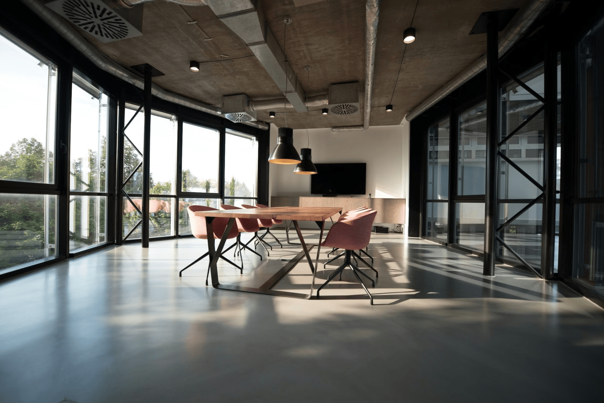 A modern conference room with large windows, a long wooden table, pink chairs, hanging lights, and a wall-mounted TV, filled with natural light and surrounded by industrial-style decor.