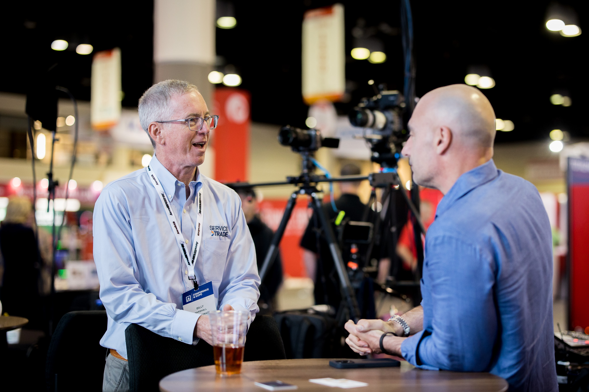 Two men have a conversation at a round table during an event or conference; one wears glasses and a name badge, the other is bald. Cameras and banners are visible in the background.