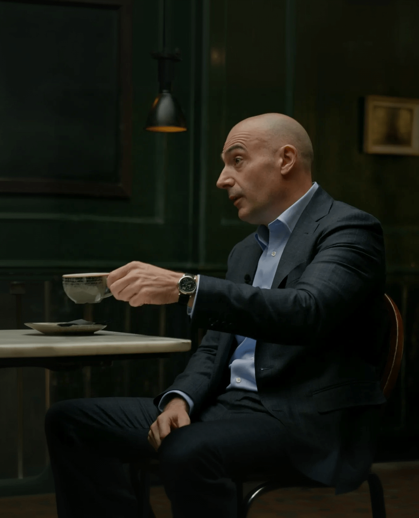 A bald man in a dark suit sits at a table holding a teacup and saucer, gesturing as he speaks in a dimly lit room with dark green walls and a hanging light above.