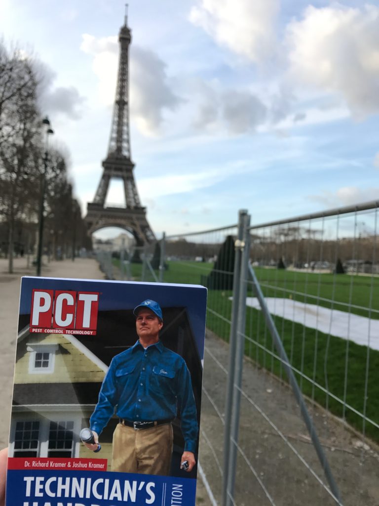 A booklet titled "Technician's Handbook" is held up in front of the Eiffel Tower in Paris. The booklet features a man in a blue uniform. The Eiffel Tower and a fenced grassy area are visible in the background.