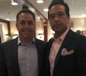 Two men wearing suits stand side by side indoors at what appears to be a formal event. The background shows soft lighting, wood paneling, and other people in the distance.