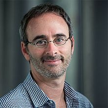 A middle-aged man with short dark hair, gray beard, and glasses, wearing a checkered shirt, smiles slightly while standing in front of a blurred, neutral background.