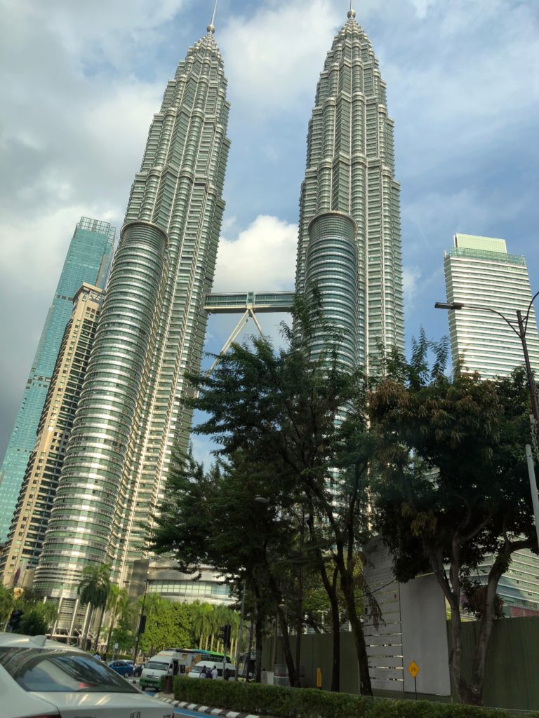 The image shows the Petronas Twin Towers in Kuala Lumpur, Malaysia, with a skybridge connecting the two skyscrapers. Trees and cars are visible in the foreground under a partly cloudy sky.