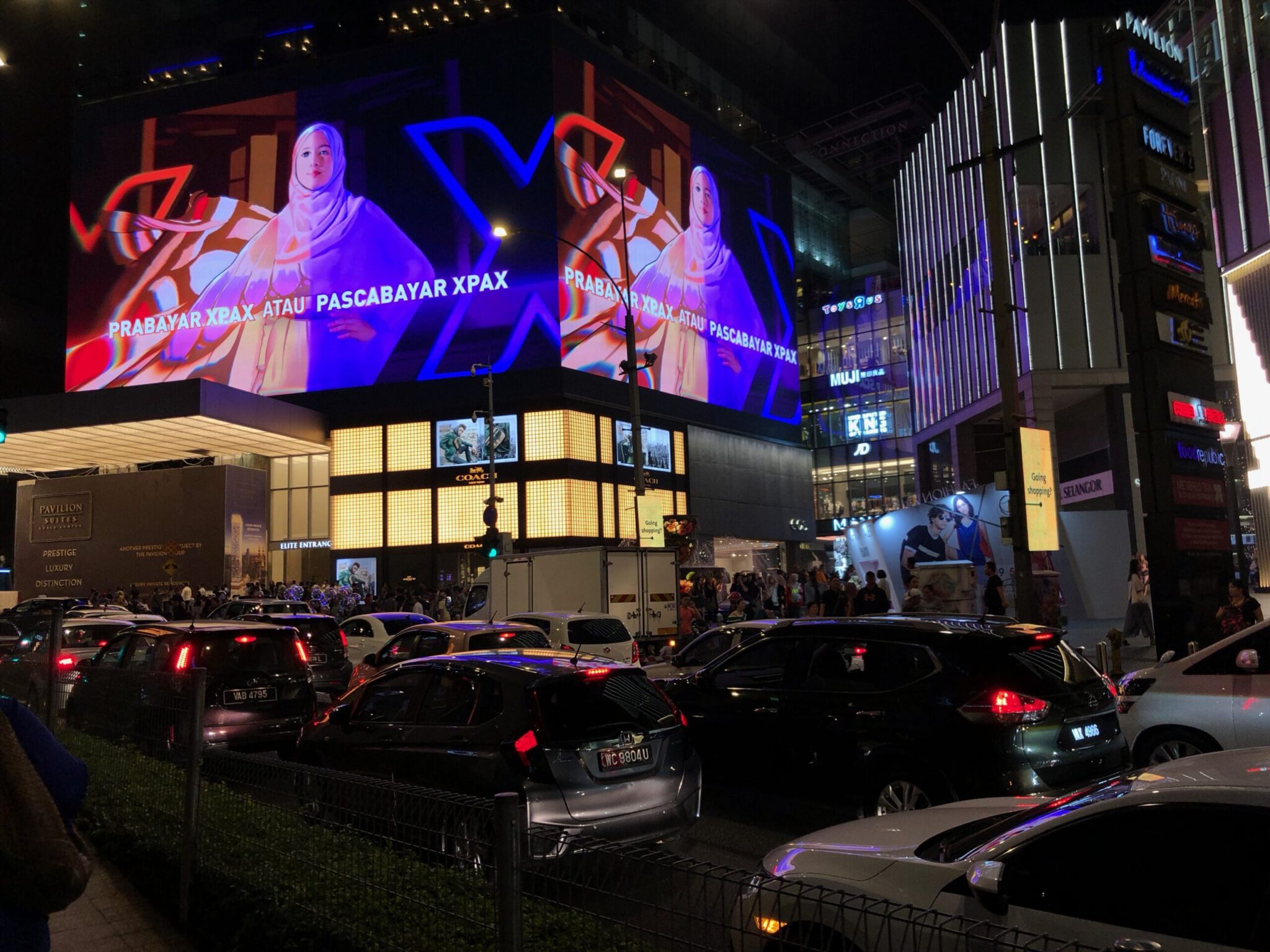 A busy city street at night with heavy traffic and illuminated buildings. A large digital billboard displays a woman in a hijab with colorful graphics and text in a foreign language. Pedestrians walk along the sidewalk.