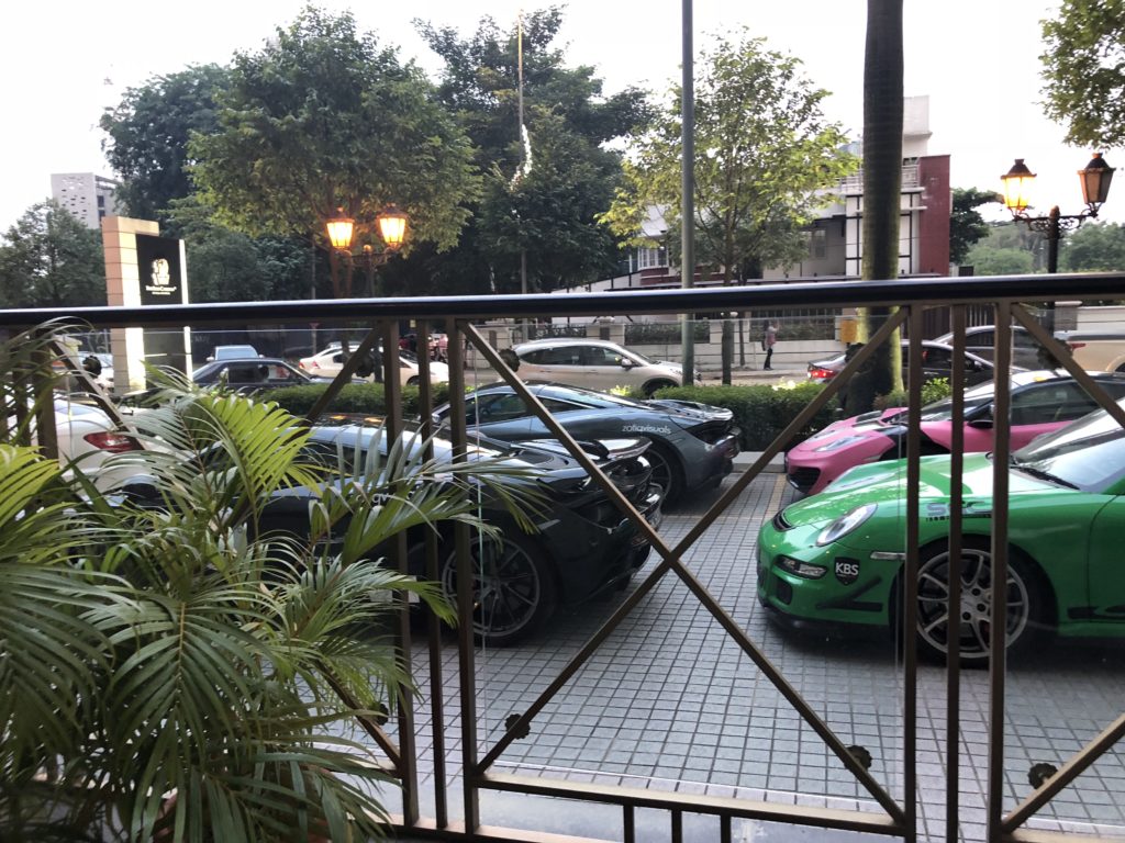Several luxury cars, including a green sports car, are parked along a tiled area outside, with plants and a metal railing in the foreground and trees, streetlights, and buildings in the background.