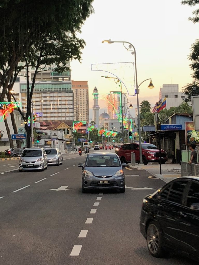 A city street at dusk with cars driving, colorful lights strung along lampposts, tall buildings, and a distant clock tower in the background. Trees line the road and people walk along the sidewalks.