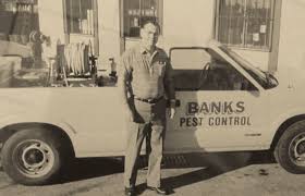 A man stands in front of a pickup truck labeled "BANKS PEST CONTROL" parked outside a building. Equipment is visible in the truck bed. The photo is in black and white.