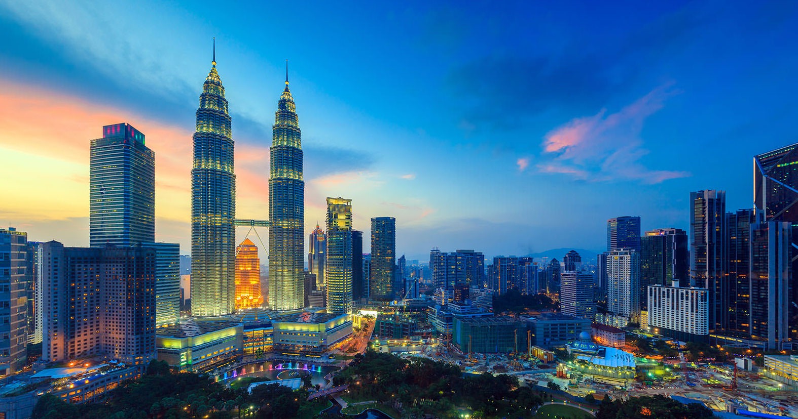 Kuala Lumpur city skyline at sunset, featuring the illuminated Petronas Twin Towers and surrounding high-rise buildings under a vibrant blue and orange sky.