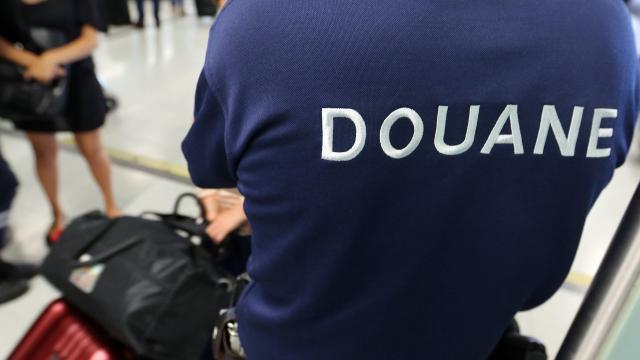 A customs officer wearing a blue shirt labeled "DOUANE" inspects luggage at an airport; other travelers and suitcases are visible in the background.