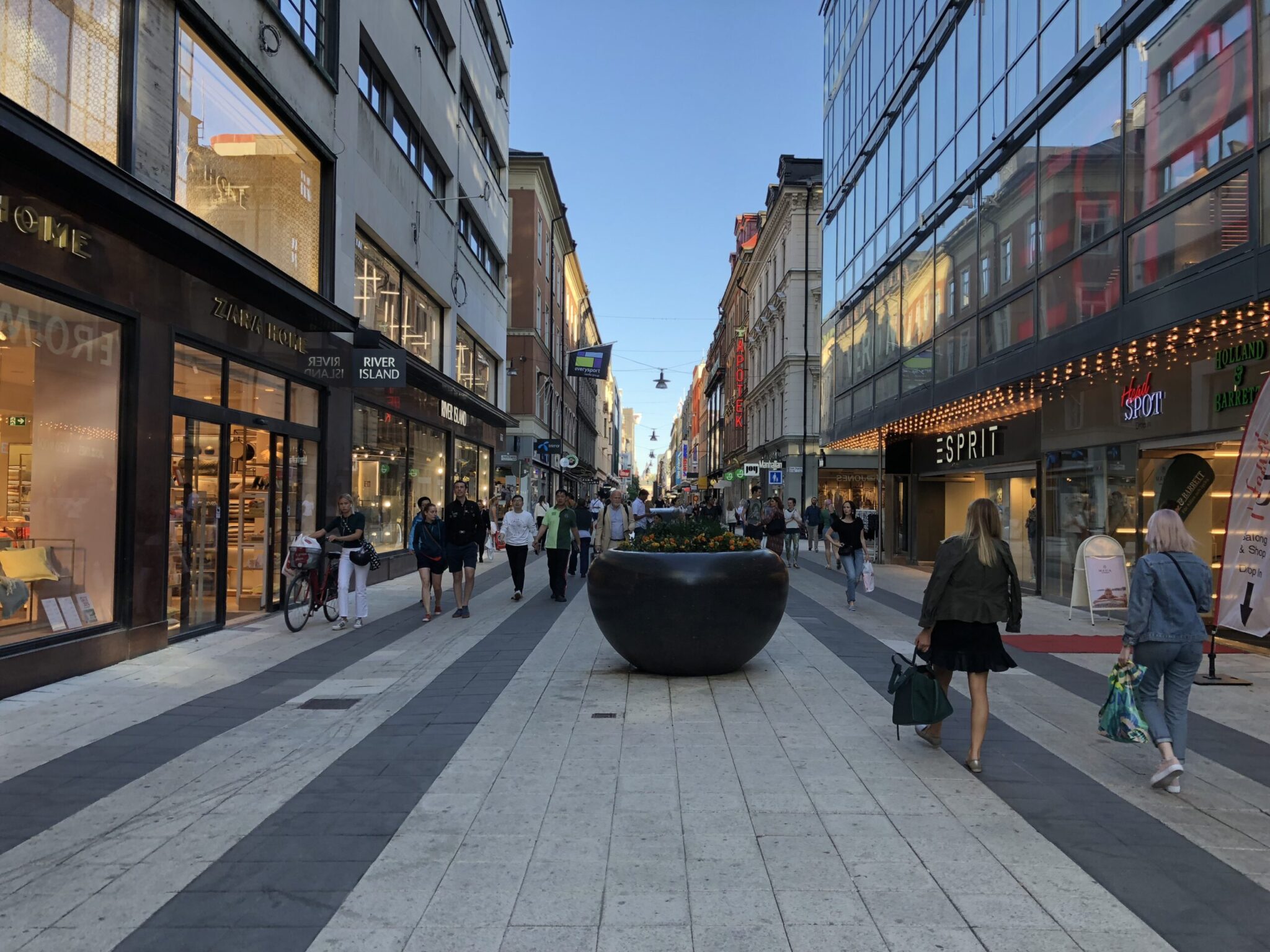 A busy pedestrian shopping street lined with stores and glass buildings, people walking and shopping, with a large round planter filled with flowers in the center of the walkway.
