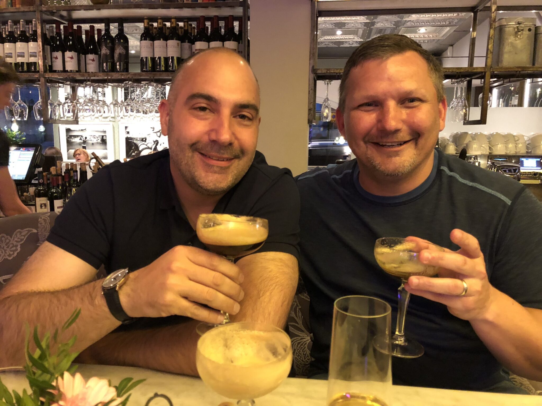 Two men smiling and raising cocktail glasses while seated at a bar, with shelves of wine bottles and glasses in the background. They appear to be enjoying a relaxed evening together.
