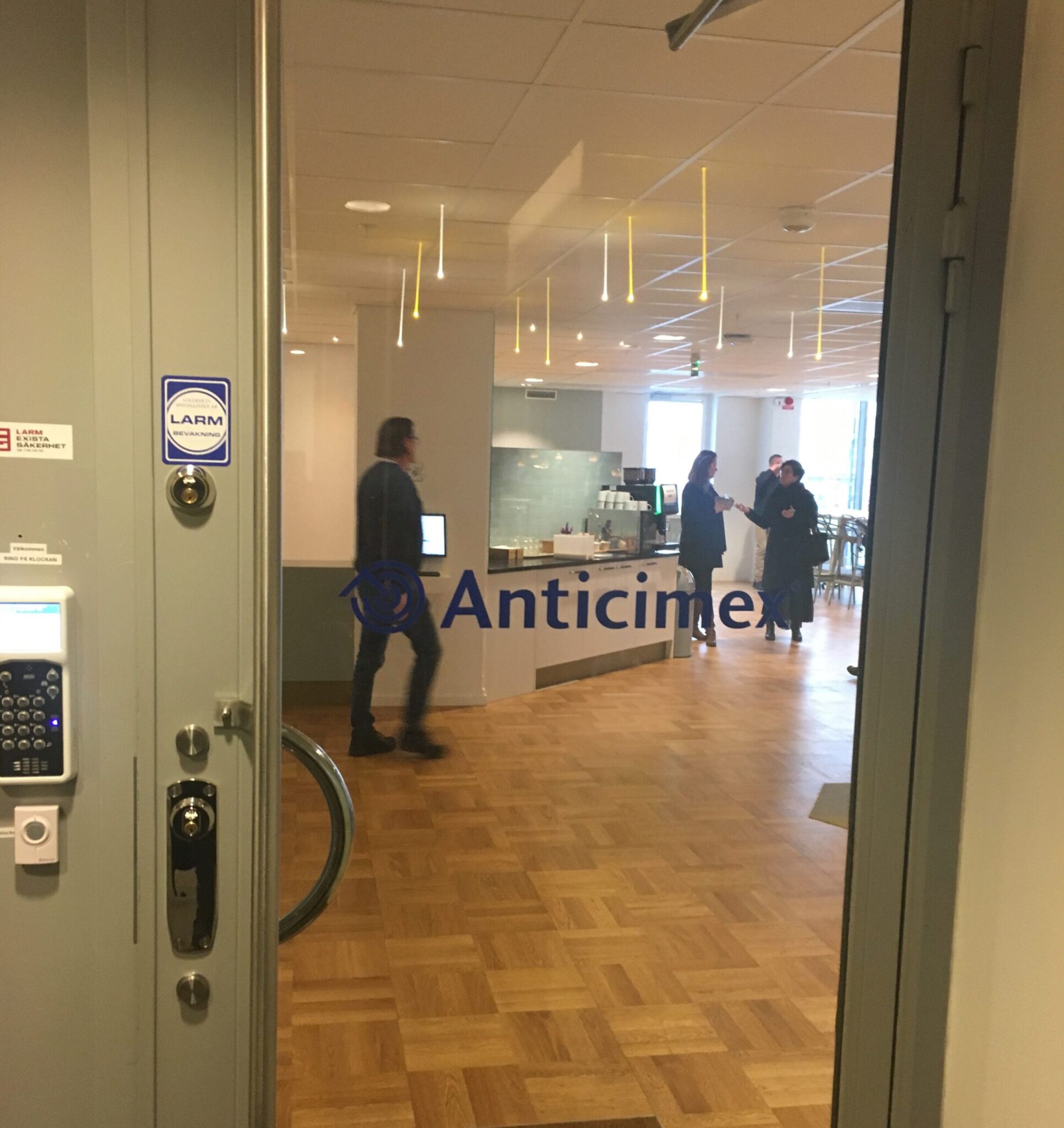 View through a glass door into an office with wooden floors, a few people standing and talking, some walking. The word “Anticimex” is printed on the glass. Ceiling lights and desks are visible inside.