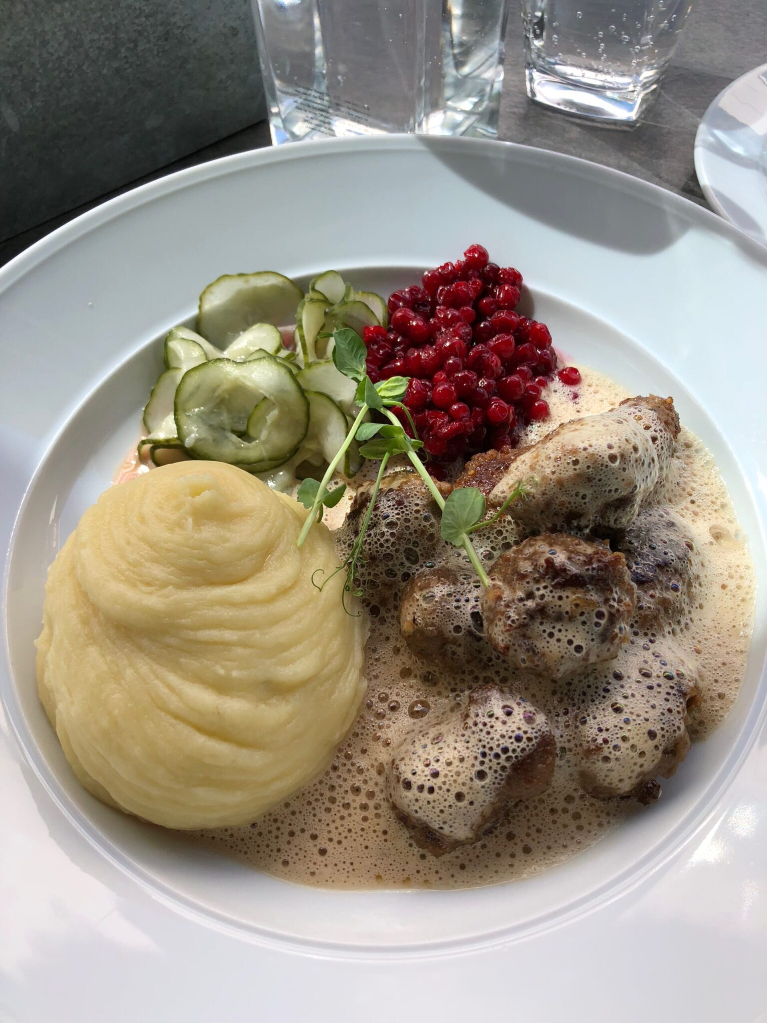 A white plate with mashed potatoes, Swedish meatballs in cream sauce, sliced pickled cucumbers, and a portion of red lingonberries. Glasses of water are in the background.