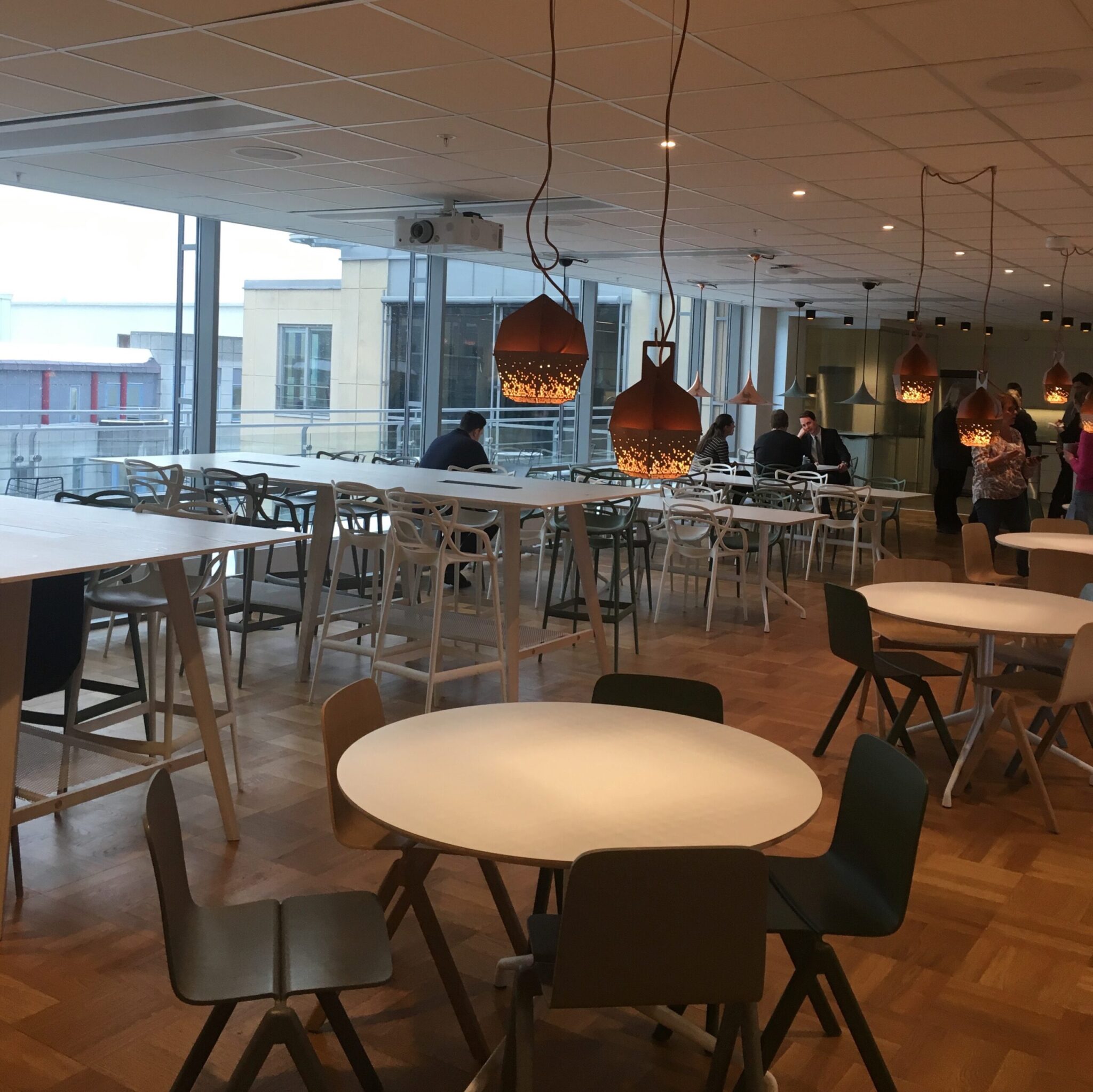 A modern cafeteria with round and rectangular tables, mixed chairs, wooden floor, and hanging copper pendant lights. People are seated and working near large windows with an urban view.