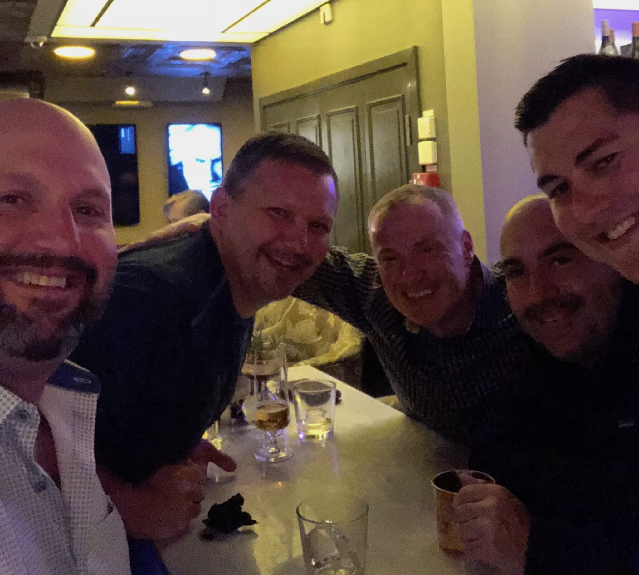 Five men are smiling and posing together at a bar table with drinks in front of them. The setting is dimly lit and casual, with background lights and a TV visible behind them.