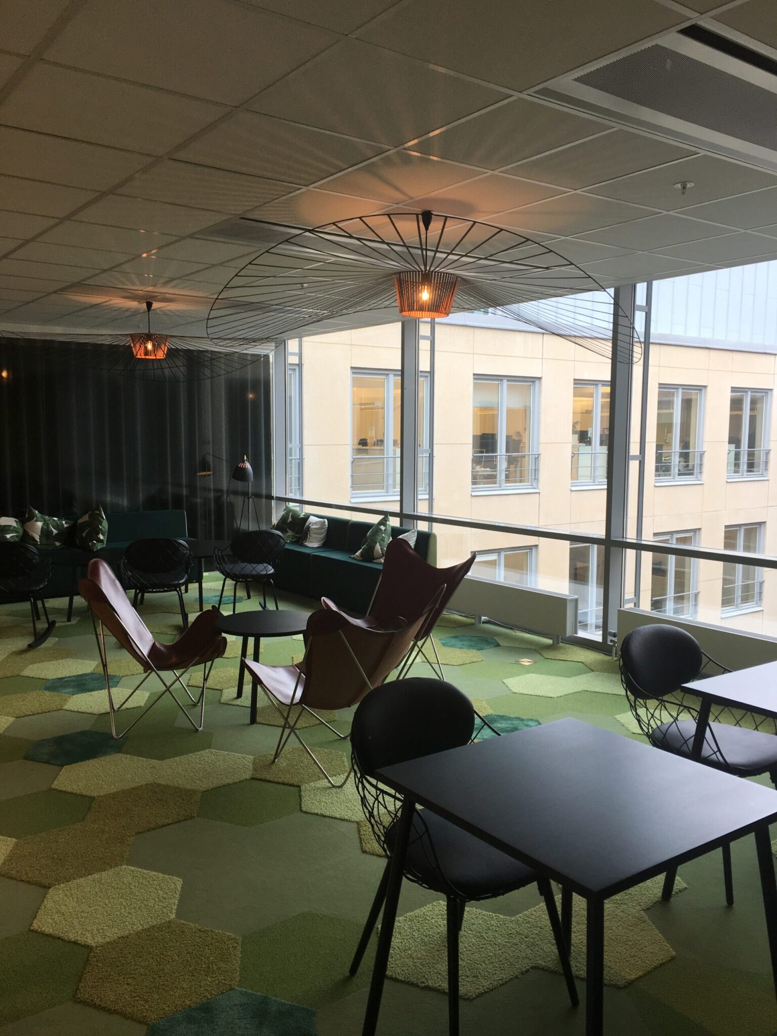 Modern office lounge with large windows, black chairs, small tables, lounge seating, and geometric green carpet tiles. Ceiling lights with wire shades hang above, and a building is visible outside the windows.