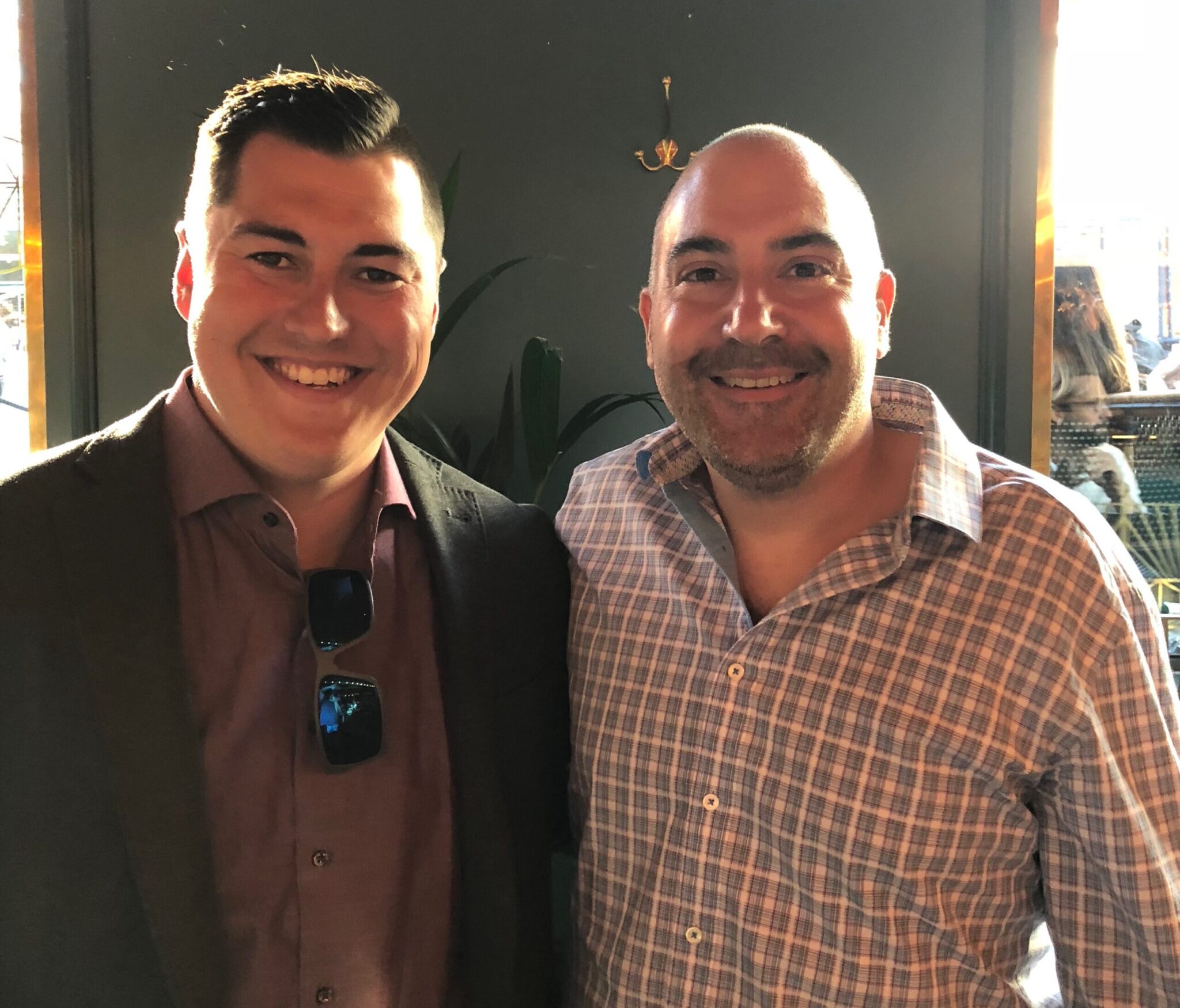 Two smiling men posing together indoors. One is wearing a dark blazer and maroon shirt, the other is in a checked button-down shirt. Both are standing in a warmly lit room with a plant in the background.