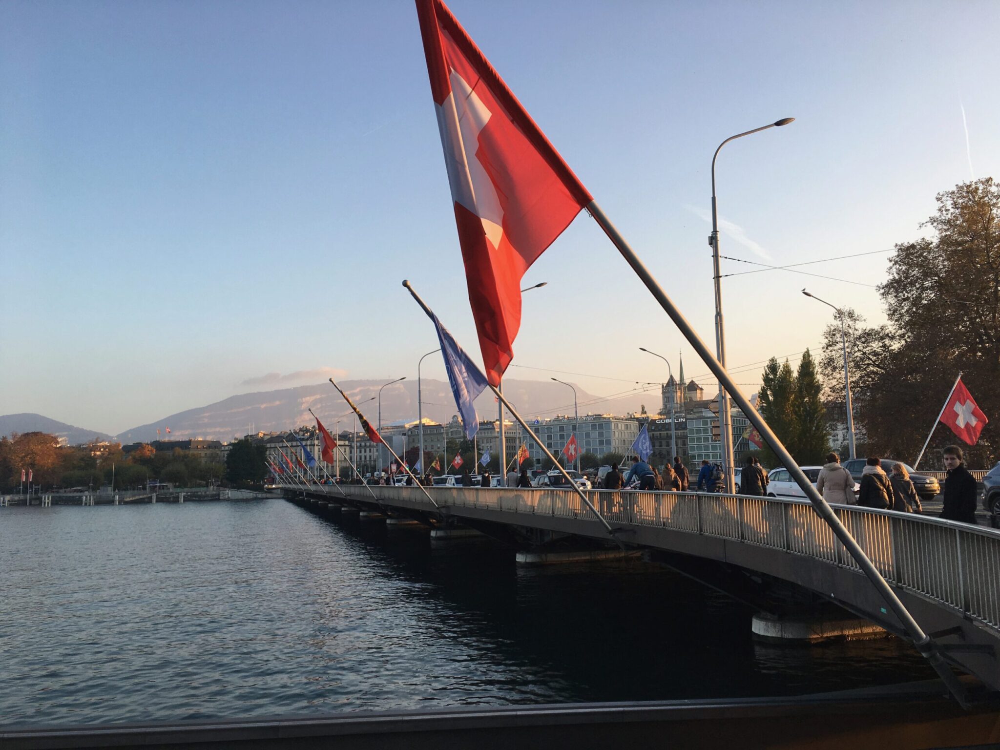 A bridge lined with Swiss flags crosses over a body of water at sunset, with people walking and city buildings in the background. Trees and mountains are visible in the distance.