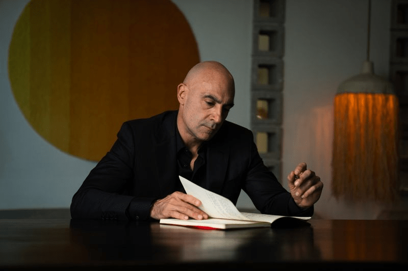 A bald man in a dark suit sits at a table, reading an open book. The background features a large yellow-orange circle on the wall and a lamp with a fringe shade, casting a warm light.