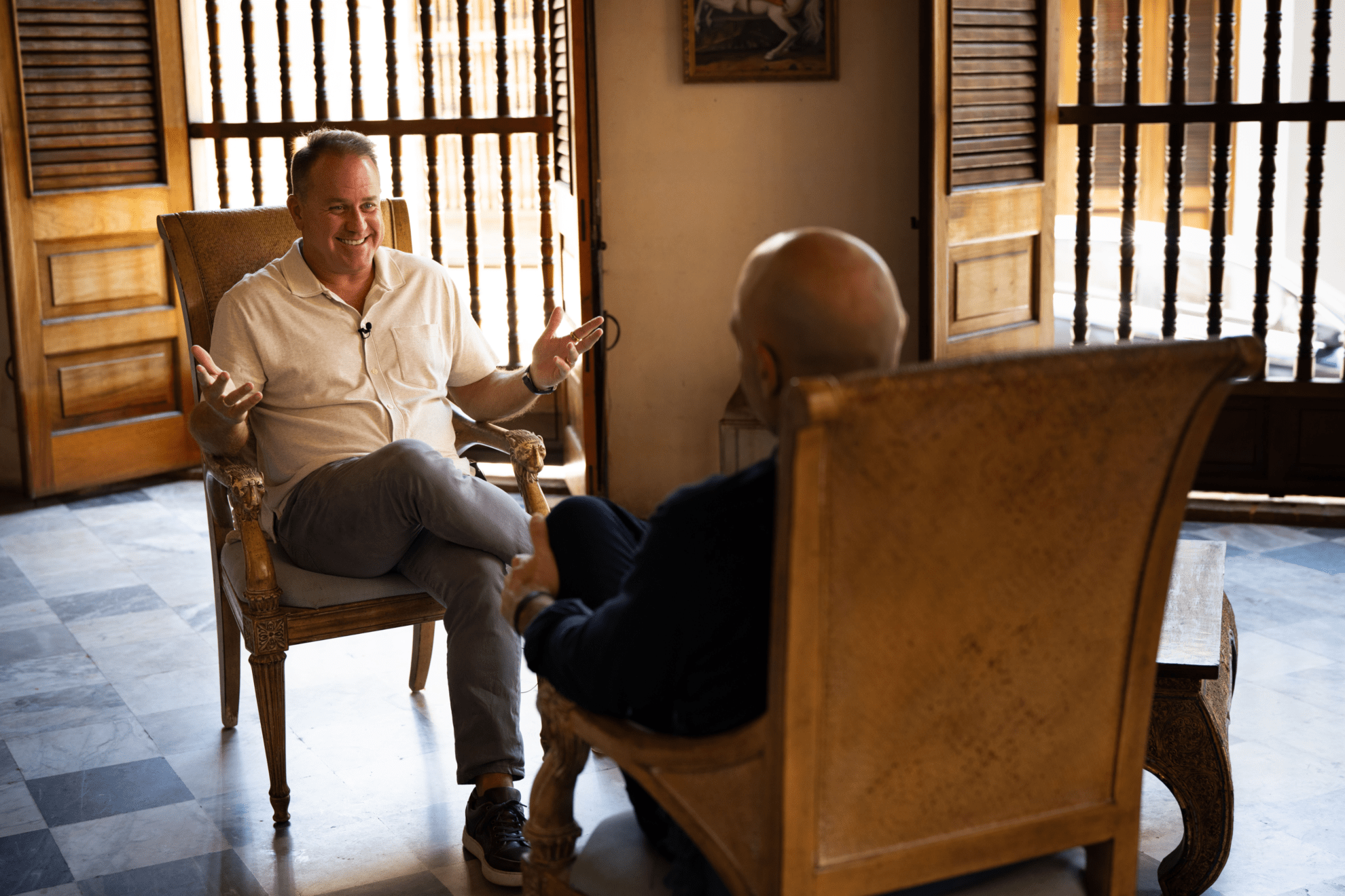 Two people sit facing each other in wooden chairs, engaged in conversation inside a sunlit room with open wooden shutters and tiled floor; one person gestures animatedly while the other listens.
