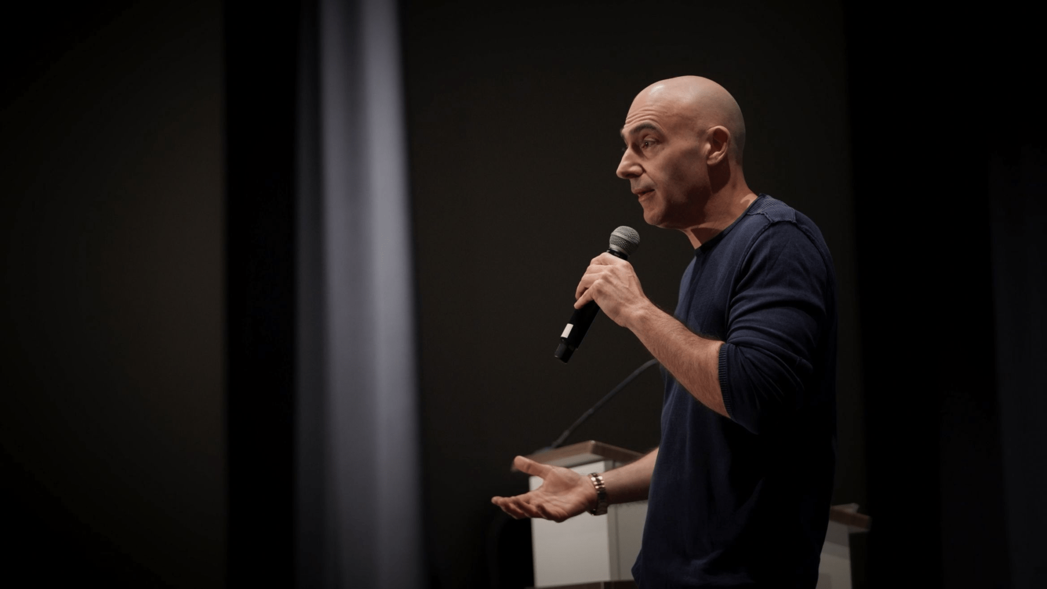 A bald man in a dark long-sleeve shirt holds a microphone and gestures while speaking on stage in a dimly lit room.