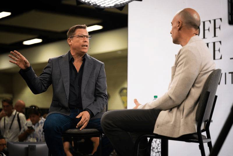 Two men sit on high stools in conversation at a public event; one gestures while speaking, the other listens. People and cameras are visible in the background, and bright lights illuminate the scene.