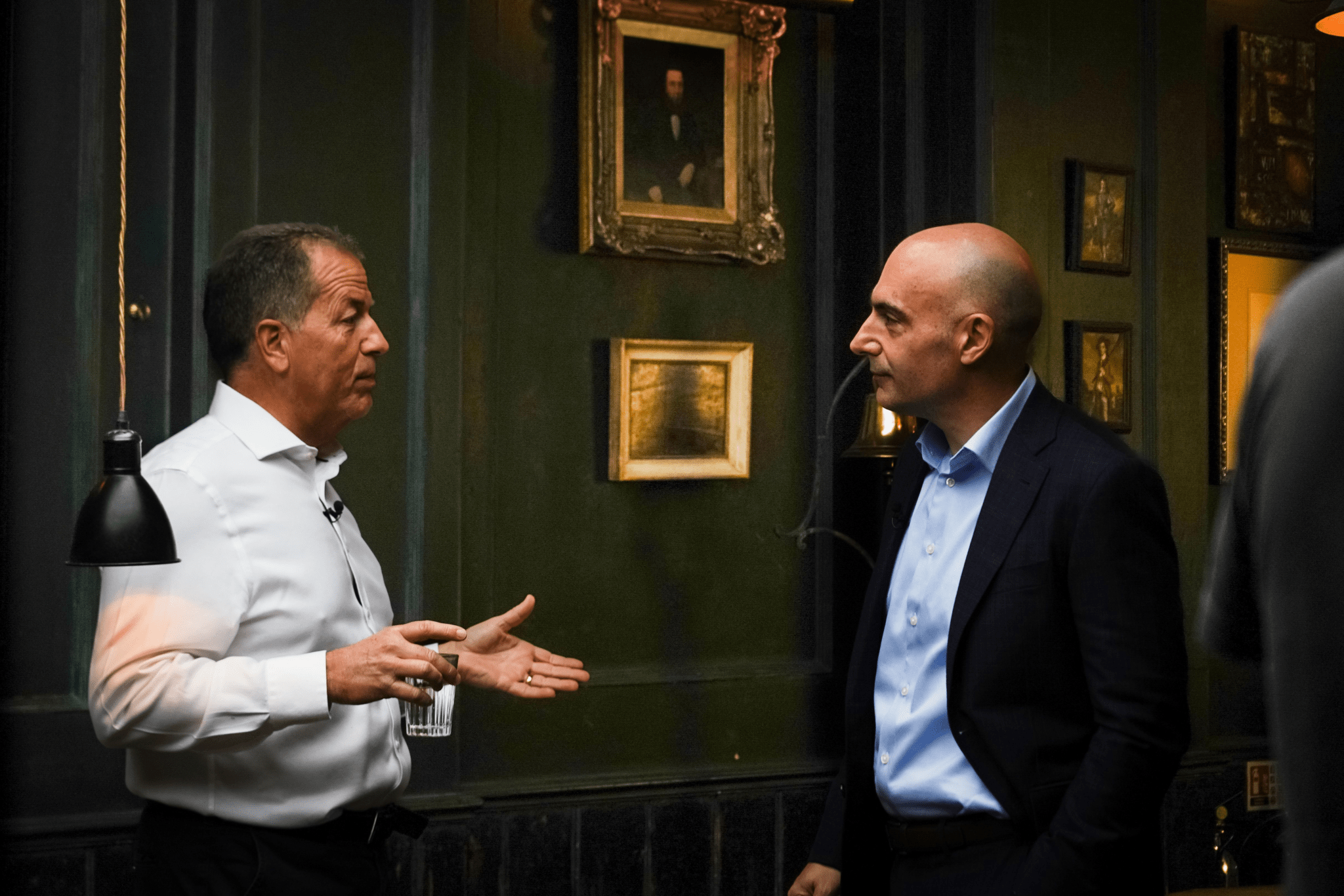 Two men in suits have a serious conversation in a dimly lit room with dark green walls, framed pictures, and a hanging lamp. One man gestures while holding a glass of water.