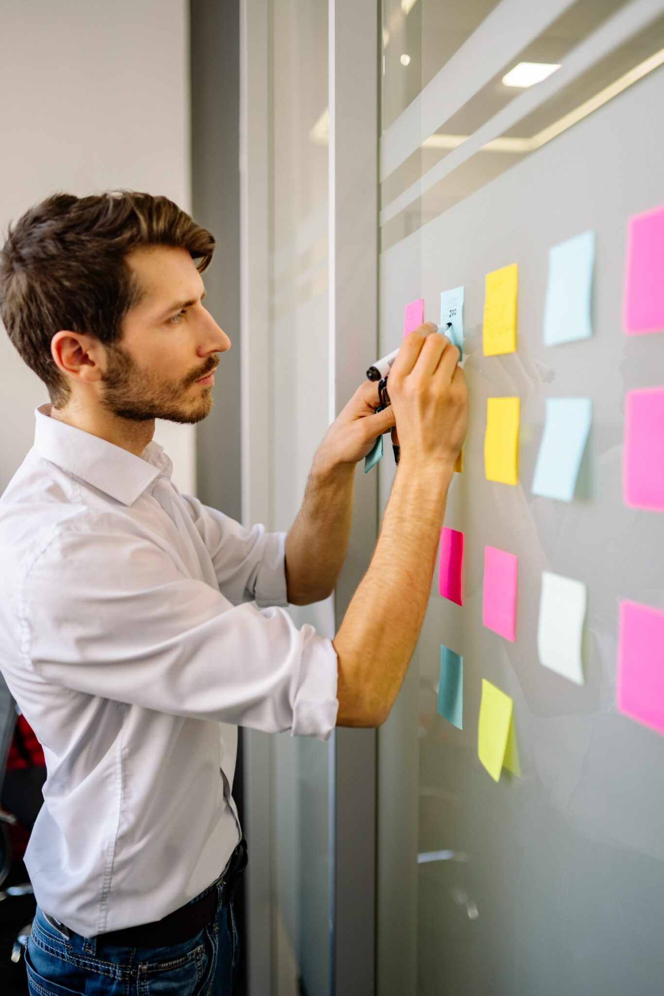 A man in a white shirt writes on a blue sticky note attached to a glass wall covered with colorful sticky notes in an office setting.