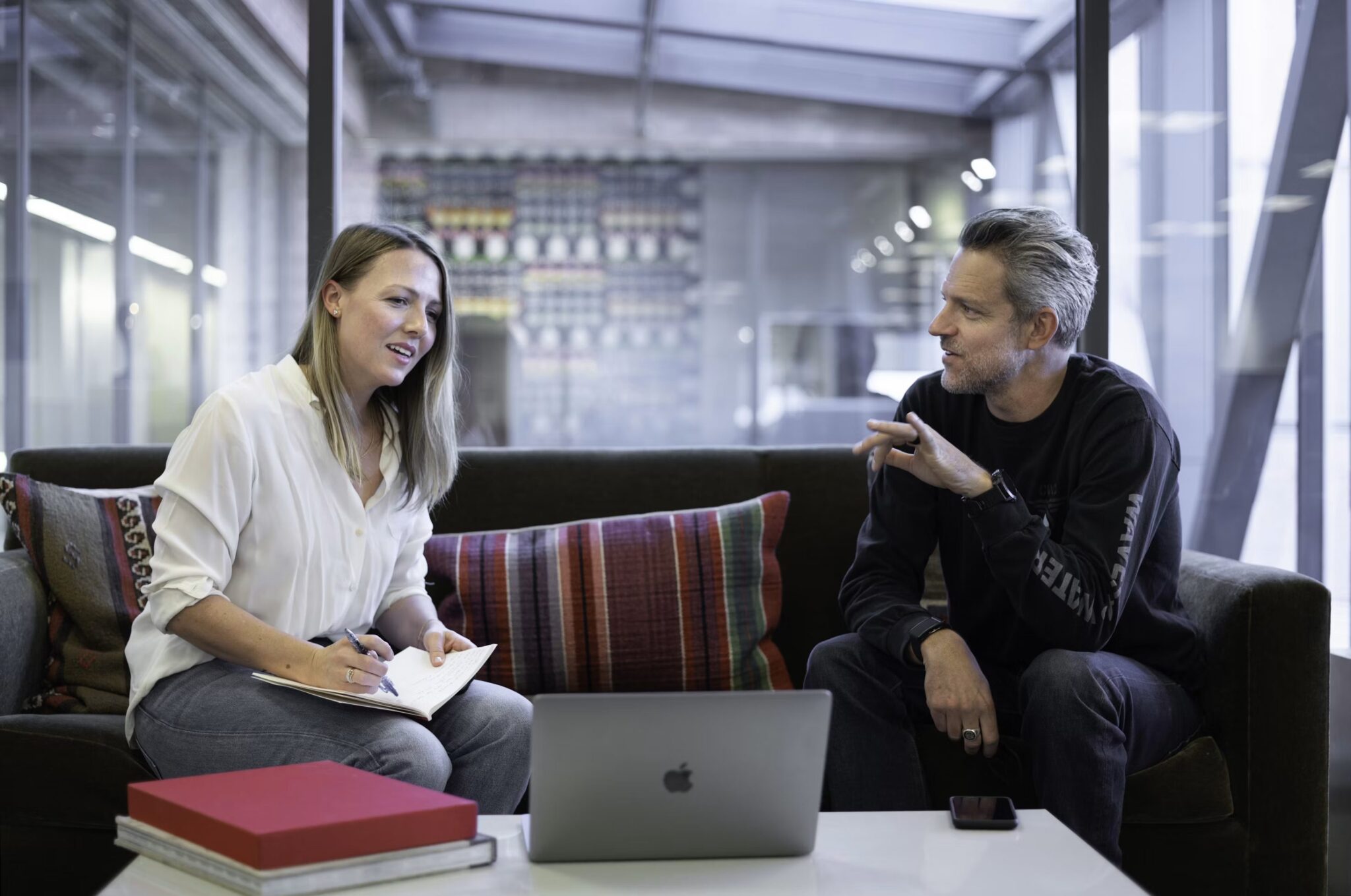 Two people sit on a sofa in a modern office, talking. The woman holds a notebook and pen, while the man gestures. An open laptop, phone, and books are on the coffee table in front of them.