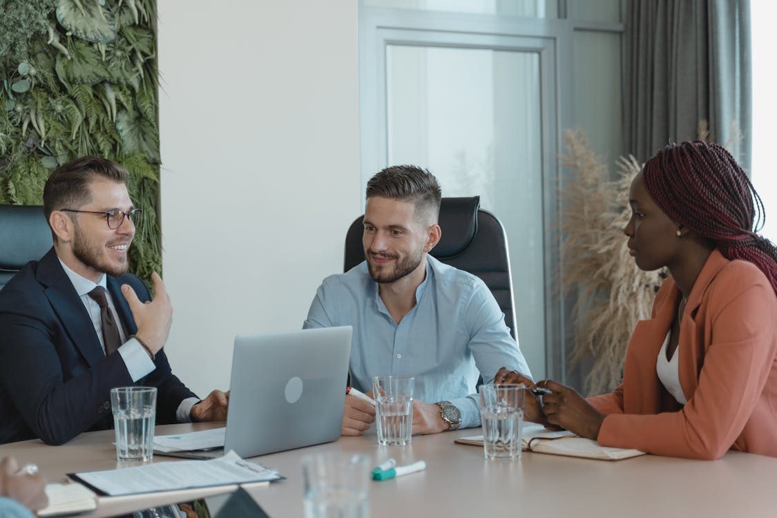 Three people sit at a conference table with a laptop, notepads, and glasses of water, engaged in a business meeting or discussion in a modern office setting.