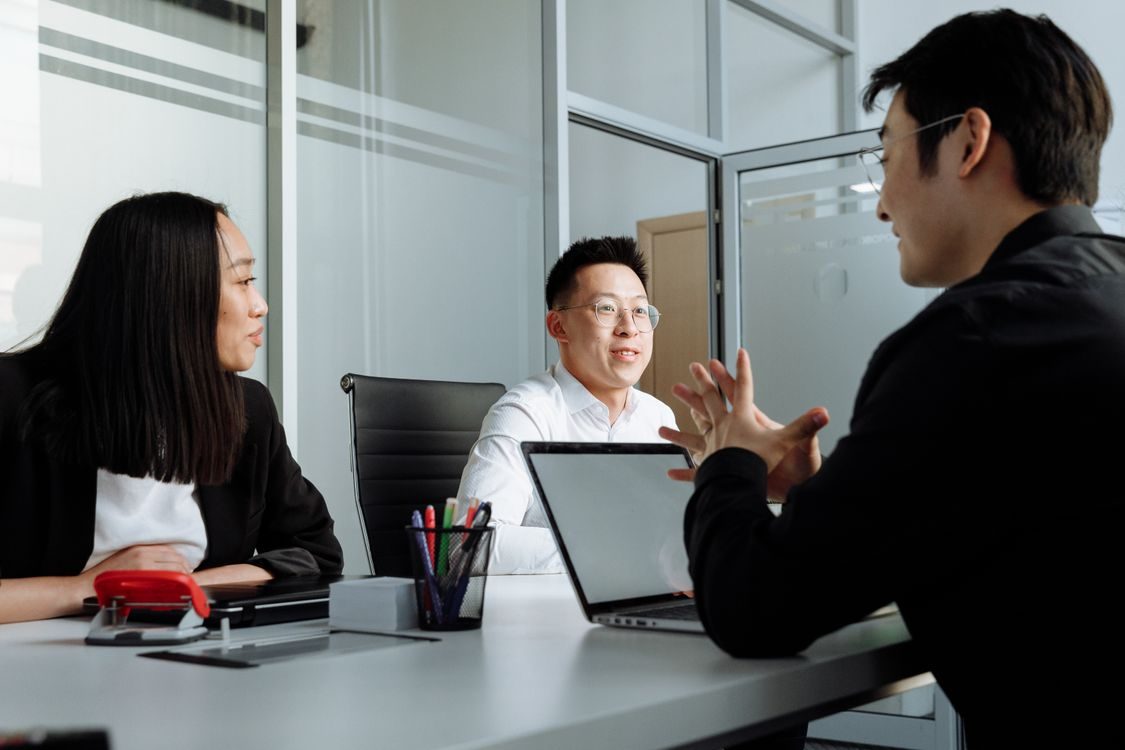 Three people sit around a desk in a modern office, engaged in conversation. Two men and one woman face each other, with laptops and office supplies on the table. Sunlight comes through glass walls in the background.