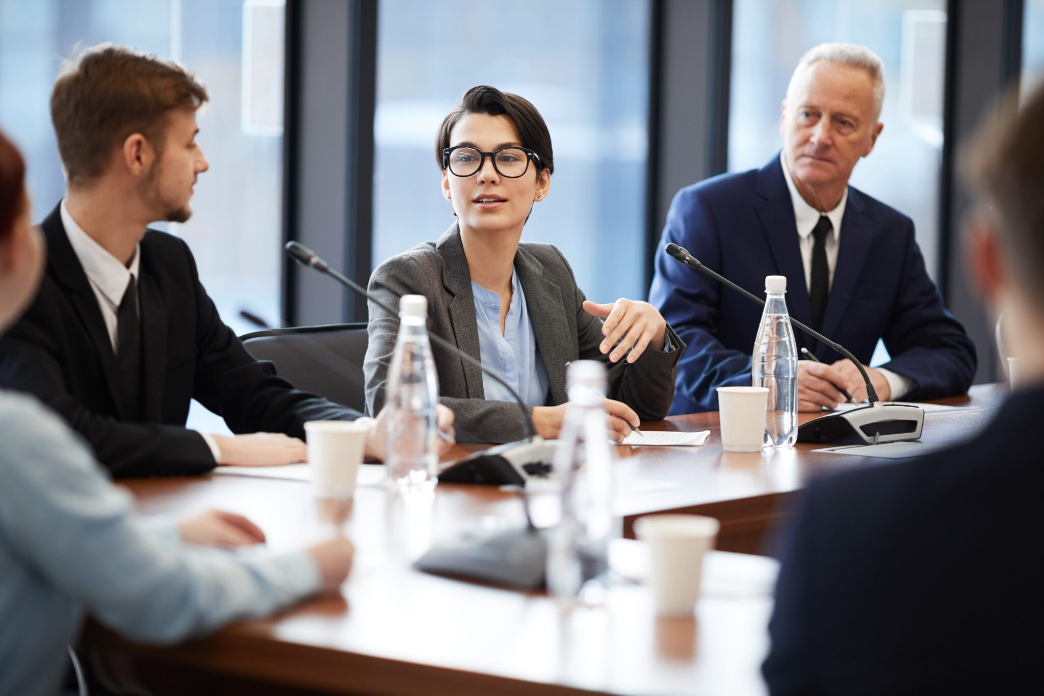 A woman in business attire speaks at a conference table with microphones, surrounded by colleagues in suits, with water bottles and papers on the table in a modern office setting.