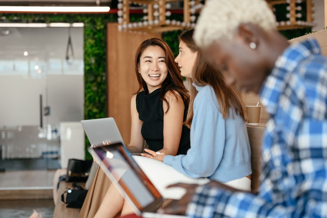 Three people are sitting indoors with laptops. Two women in the background are smiling and talking, while a person in the foreground is focused on their laptop. The setting appears to be a modern, casual workspace.