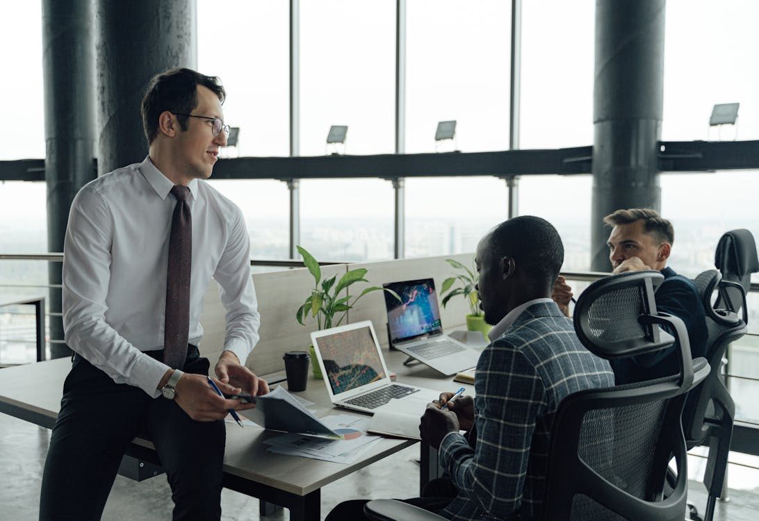 Three men in business attire have a discussion in a modern office with large windows. One stands by a desk holding papers, while the other two sit at laptops displaying charts. Plants and city views are visible in the background.