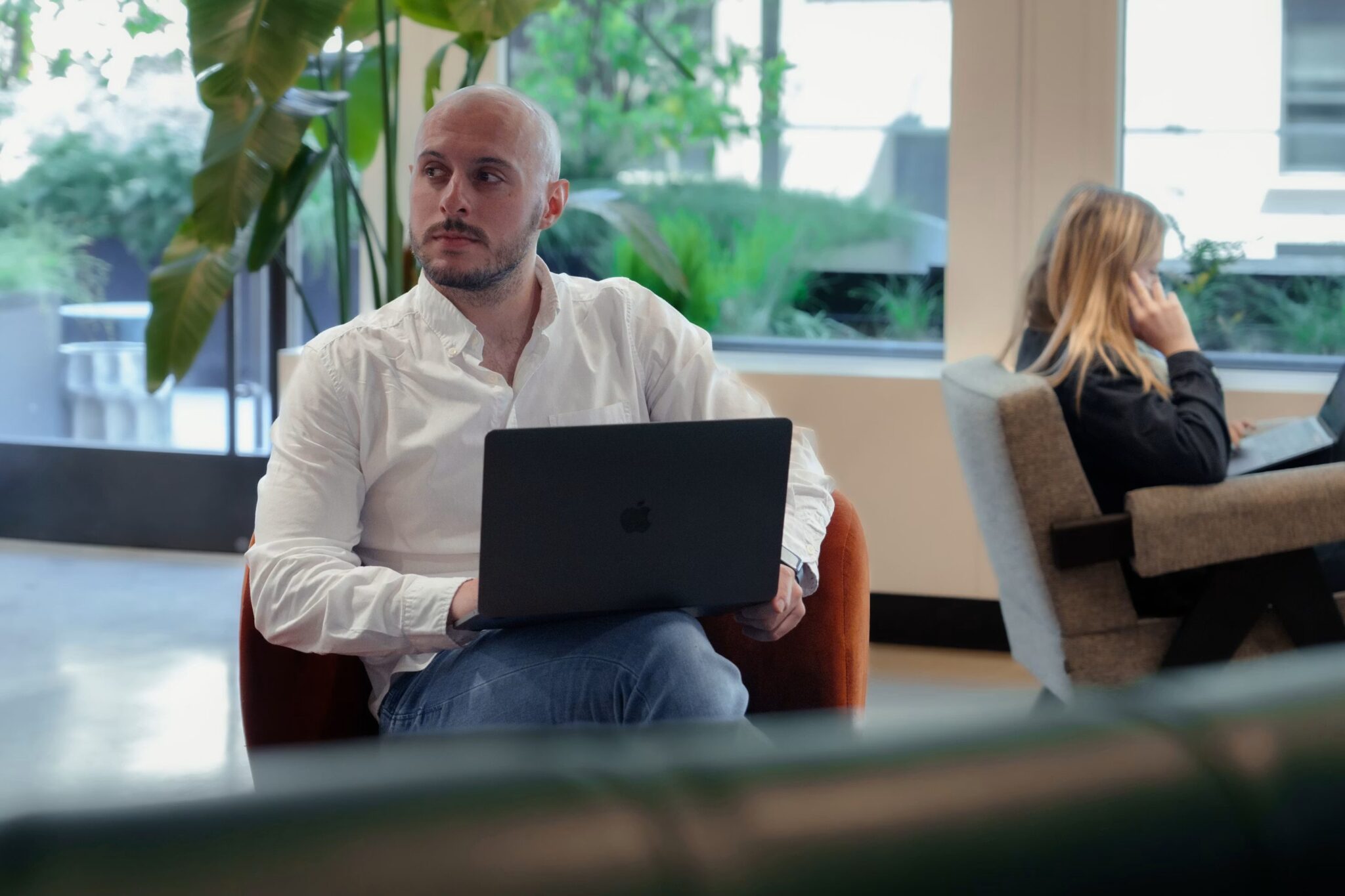 A man with a shaved head and a white shirt sits in a modern office lounge using a laptop, looking to his left. In the background, a woman is sitting on a chair, talking on the phone.