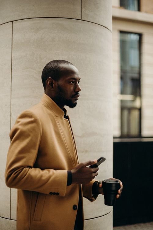 A man in a tan coat stands outdoors by a large stone column, holding a smartphone in one hand and a reusable coffee cup in the other, looking ahead with a focused expression.