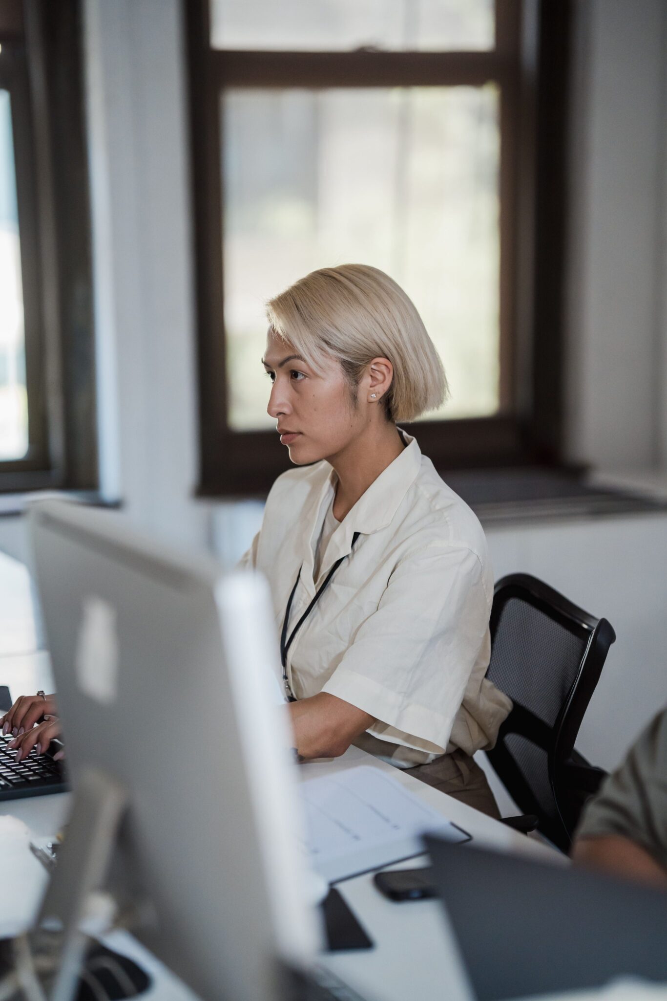 A person with short blonde hair sits at a desk, working on a computer in a bright office with large windows in the background. Papers and office supplies are visible on the desk.