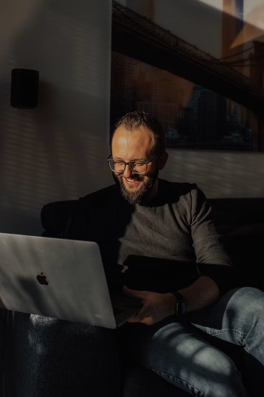 A man with glasses and a beard, wearing a dark sweater, sits on a couch using a MacBook laptop. Sunlight casts shadows across his face and the wall behind him. He appears to be smiling.