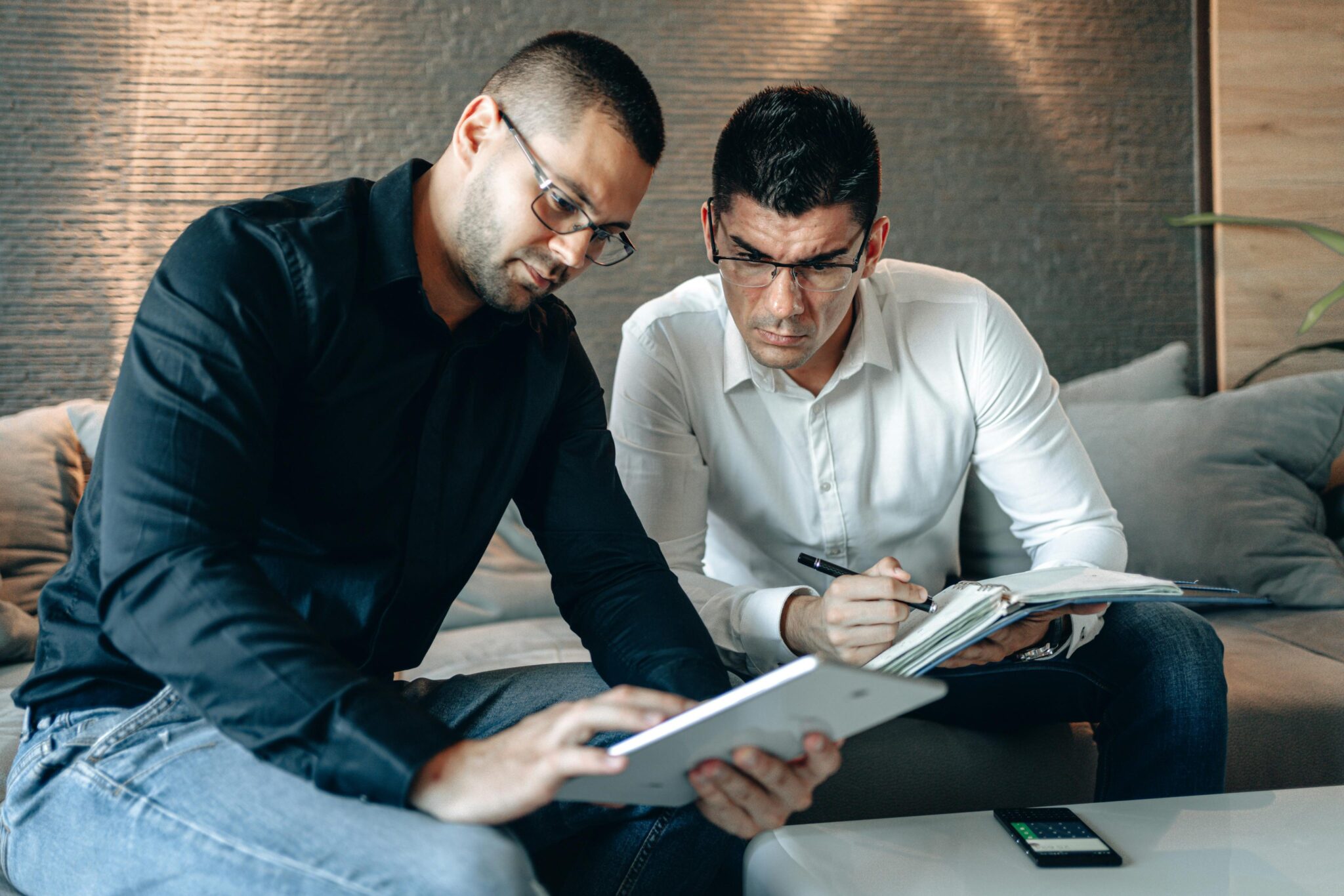 Two men sitting on a couch, one in a black shirt holding a tablet, the other in a white shirt writing in a notebook, both focused on their work. A calculator is on the table in front of them.