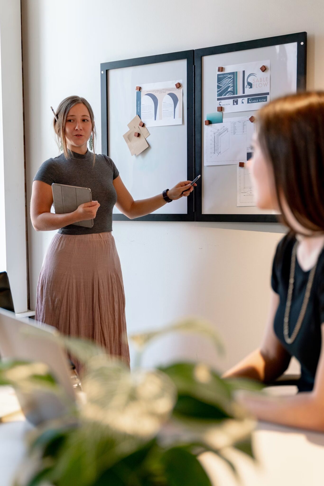 A woman stands and presents information on a bulletin board to a seated colleague in an office setting. Papers, diagrams, and notes are pinned to the board. A plant and a laptop are visible in the foreground.