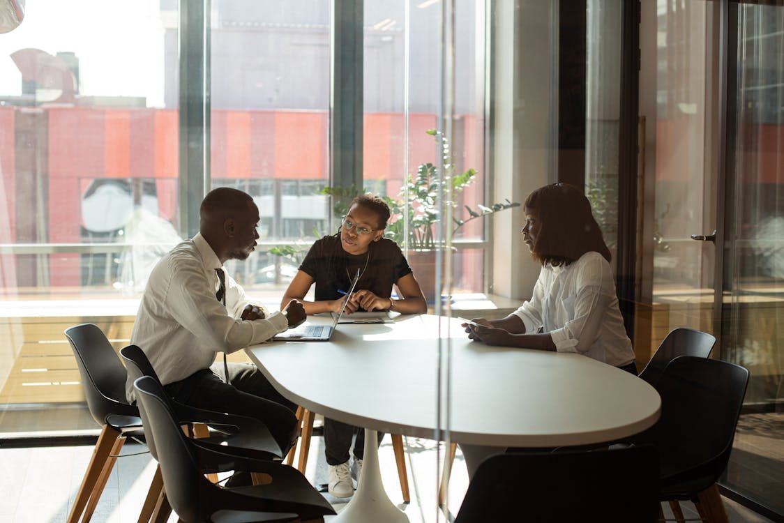 Three people are sitting around a white oval table in a modern office, having a discussion. Two women and one man appear engaged in conversation, with a laptop and notepads on the table. Large windows provide natural light.