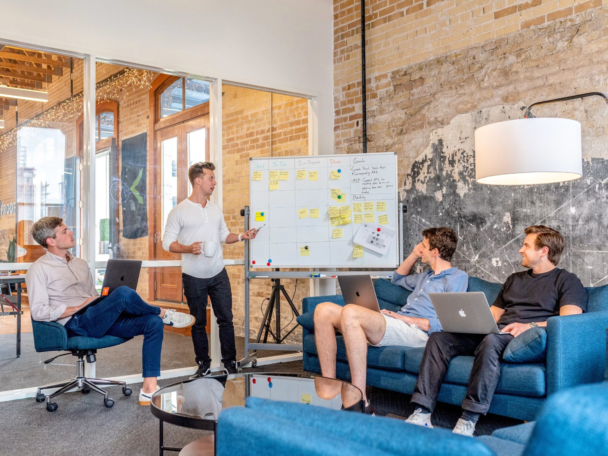 Four people are in a modern office. One person stands by a whiteboard with sticky notes, giving a presentation, while the other three sit on a sofa with laptops, appearing engaged and attentive.