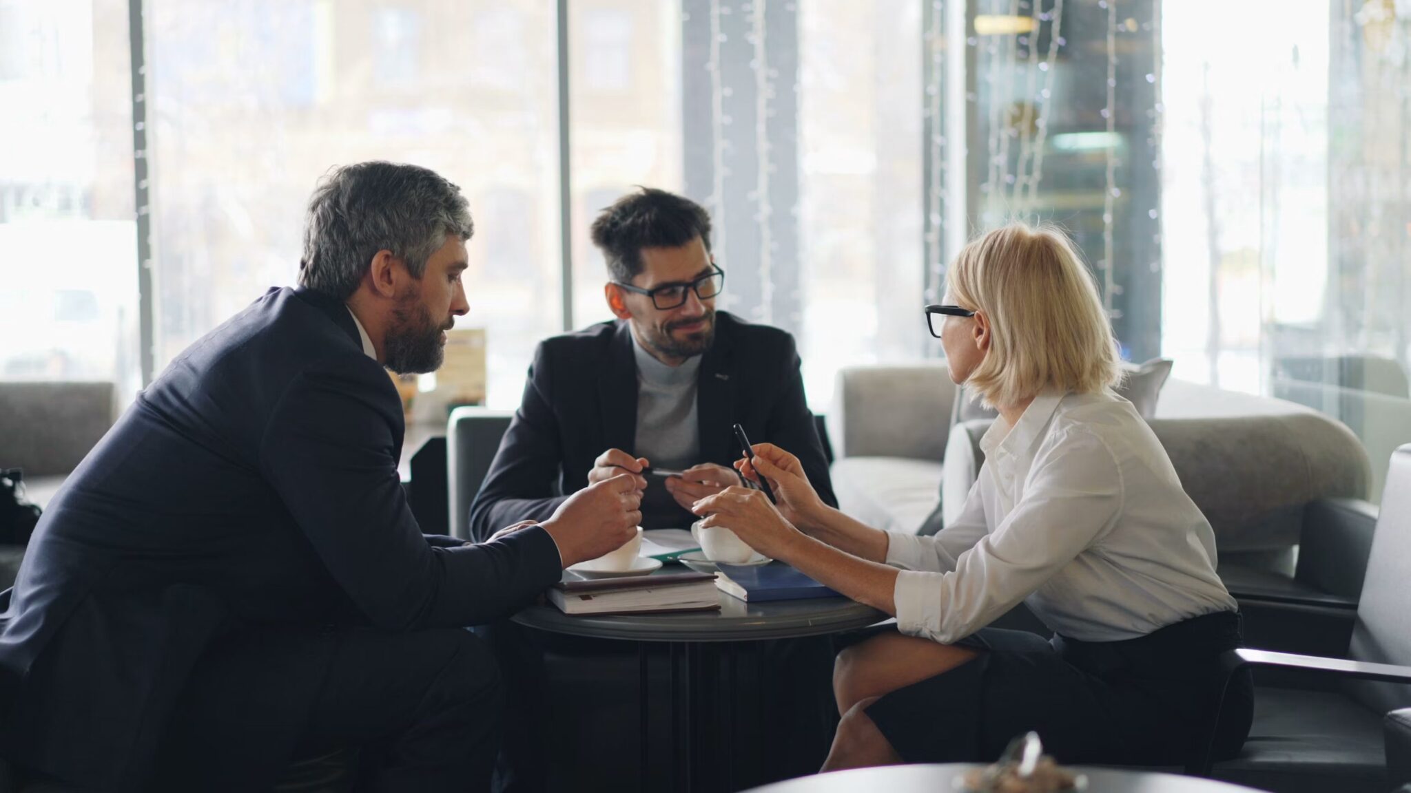 Three people in business attire sit around a table in a modern office, engaged in discussion with notebooks and coffee cups in front of them. Large windows and natural light are visible in the background.