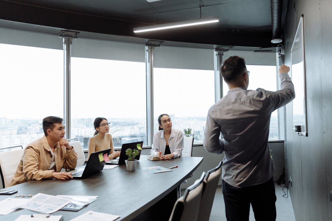 A man stands and writes on a whiteboard during a business meeting, while three colleagues seated at a conference table watch attentively with laptops and papers in front of them.