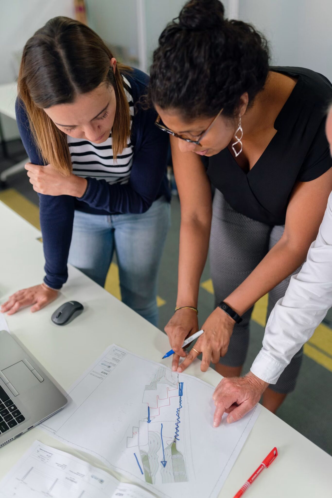 Two women and one man stand around a desk, closely examining architectural plans or diagrams. One woman points at the paper with a blue pen, while the others observe attentively. A laptop and pens are also on the desk.