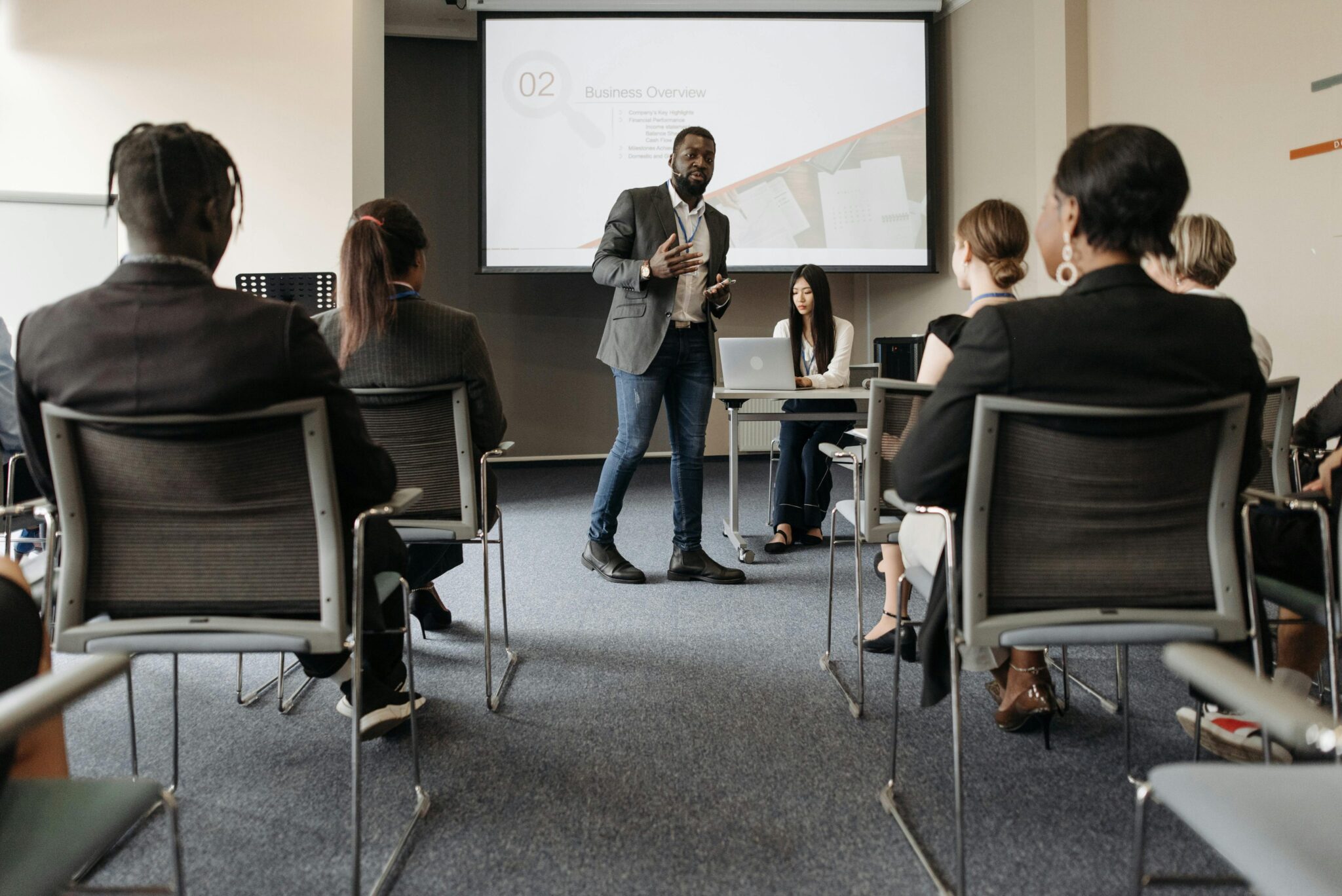 A man stands and speaks to an audience in a conference room while a woman sits at a laptop behind him. A presentation slide reading “Business Overview” is projected on a screen in the background.
