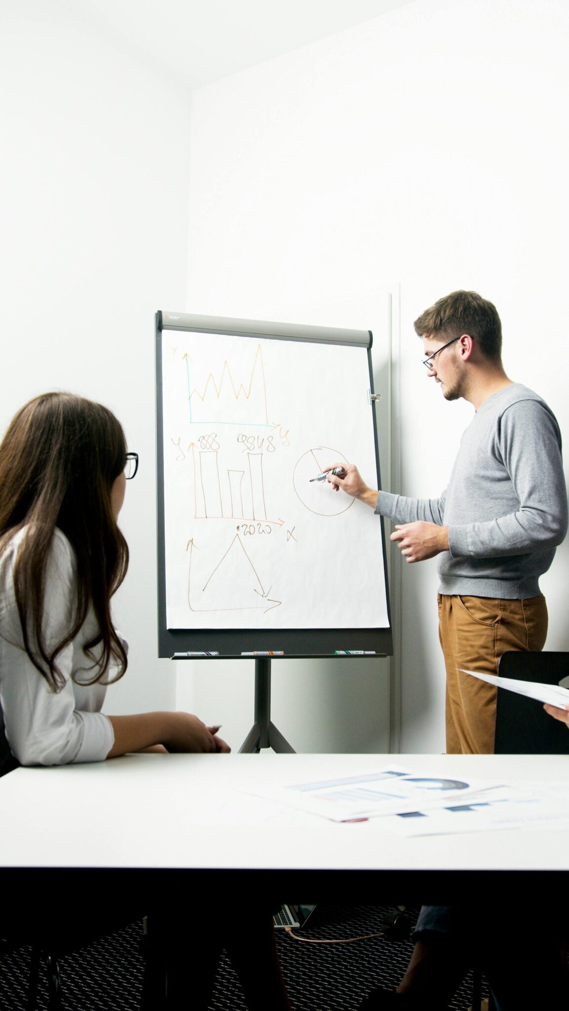 A man stands next to a flip chart with graphs and diagrams, explaining something to a seated woman in an office setting.